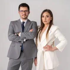 Para w garniturach, mężczyzna z rękami skrzyżowanymi na piersi, kobieta z ręką na biodrze.
