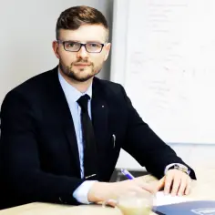 Mężczyzna w garniturze i okularach siedzi przy biurku, pisząc coś na kartce. Obok filiżanka kawy.