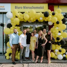 Grupa ludzi pozuje przed biurem nieruchomości "Great House" ozdobionym żółtymi balonami.