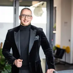 Mężczyzna w czarnym garniturze i okularach, uśmiecha się, stoi w nowoczesnym biurze.