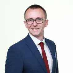 Mężczyzna w okularach, granatowej marynarce i czerwonym krawacie, uśmiecha się na białym tle.
