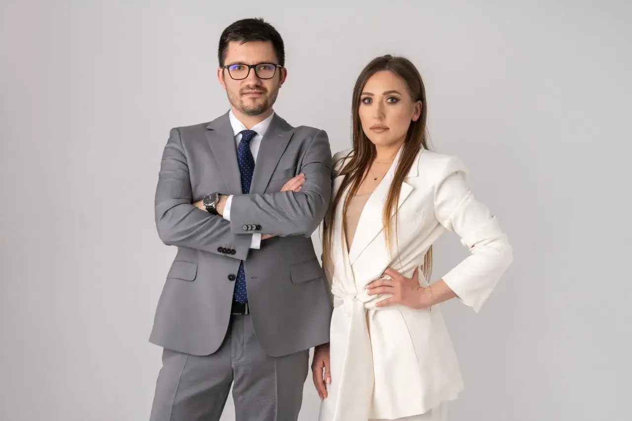 Mężczyzna w szarym garniturze i kobieta w białej marynarce stoją obok siebie na jasnym tle.