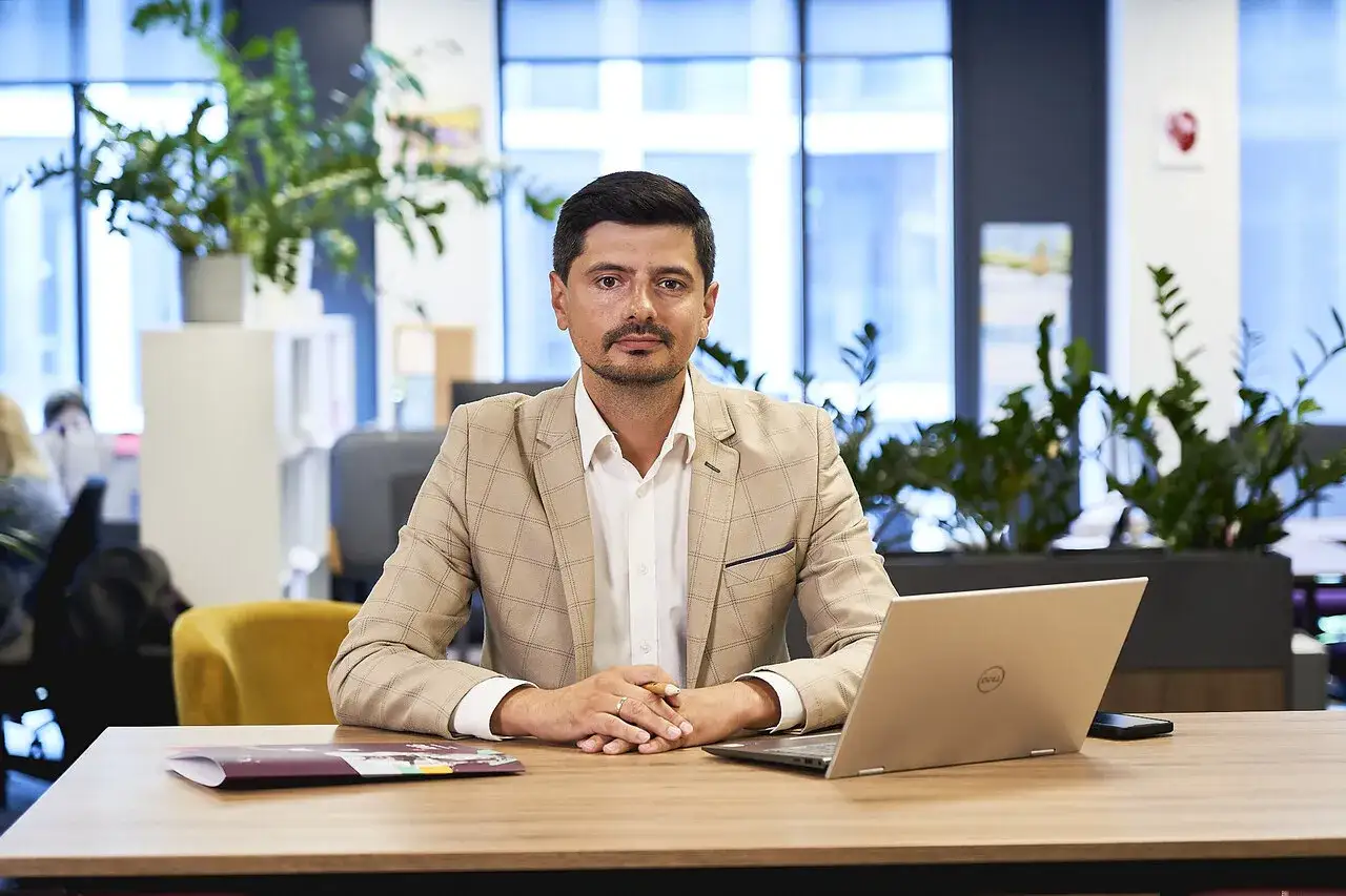 Mężczyzna w beżowej marynarce siedzi przy biurku z laptopem i dokumentami.