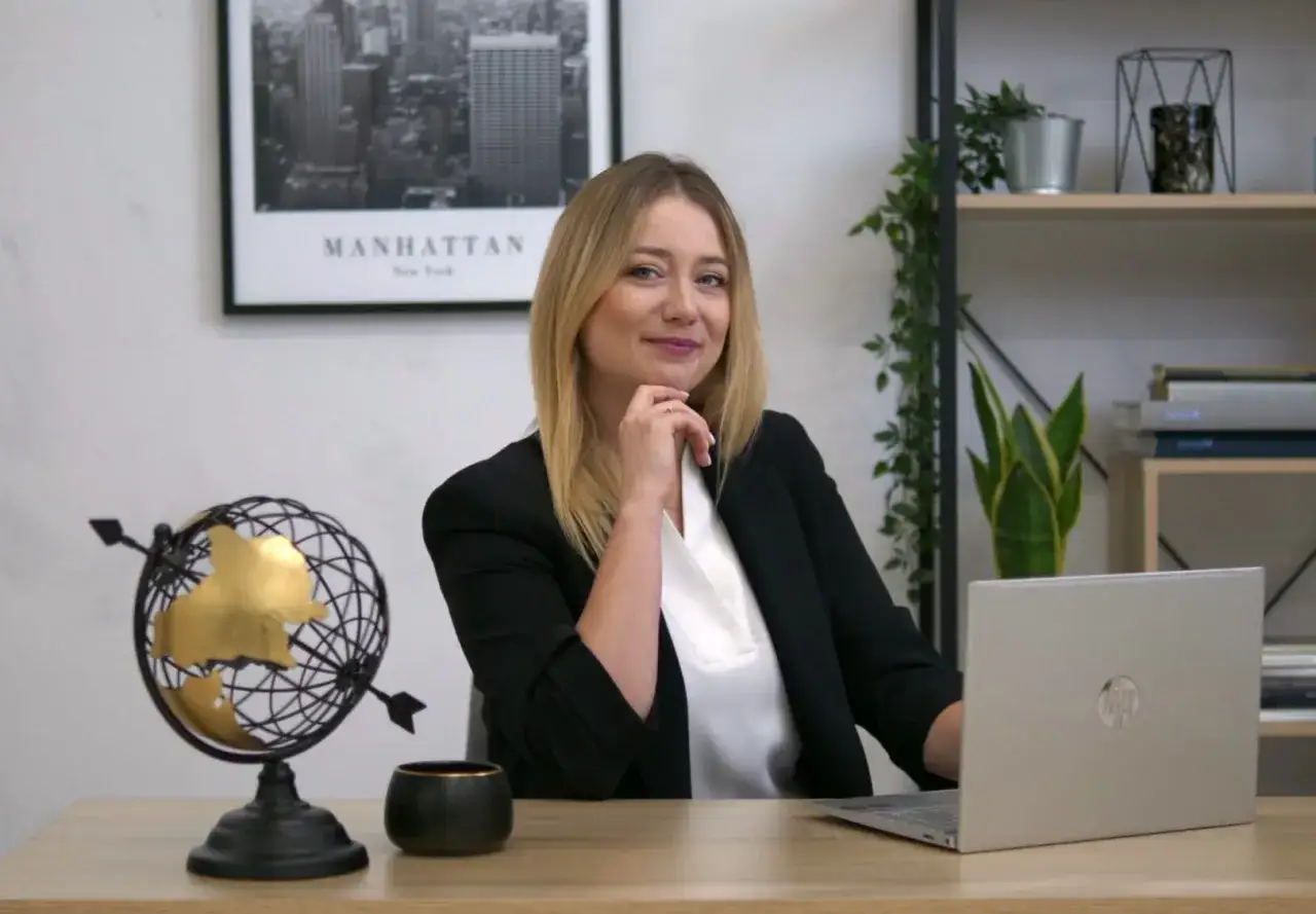 Kobieta w czarnej marynarce siedzi przy biurku z laptopem, globusem i doniczką.