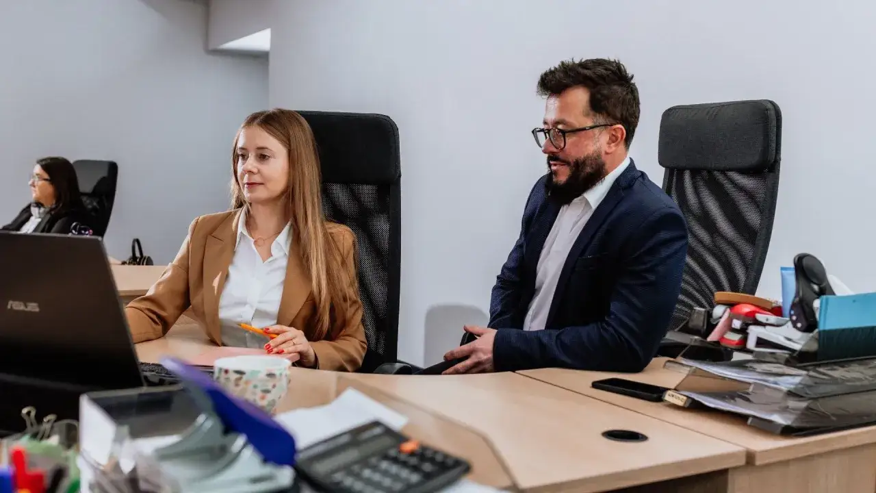 Kobieta i mężczyzna w biurze pracują przy biurku z laptopem i dokumentami.