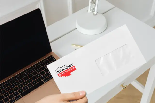 Chcesz wiedzieć, jak sprawdzić czy jestem w KRD? Na biurku leży koperta z logo Krajowego Rejestru Długów, obok laptopa.